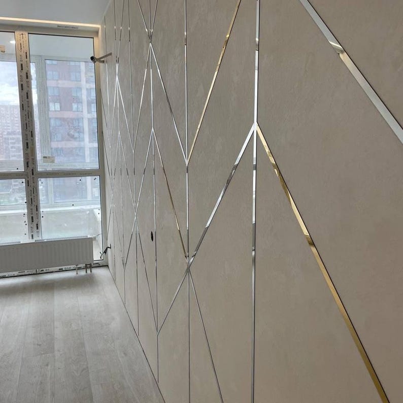 May include: A wall paneling system featuring large, matte beige rectangular panels arranged in a geometric pattern. Thin, reflective silver metal strips are inlaid between the panels, creating a continuous zigzag and diamond design across the surface.