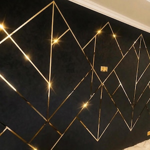 May include: A dark black wall featuring a geometric pattern of thin, reflective gold metallic strips that create intersecting triangles and diamond shapes across the surface, with bright light reflections highlighting the metallic lines.
