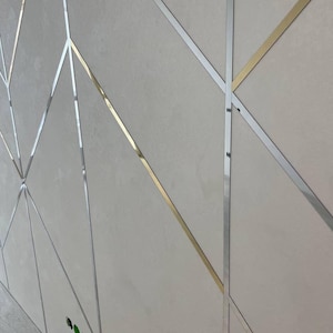 May include: A wall panel featuring a light gray, matte-textured surface divided by a geometric pattern of thin, intersecting metal strips. The strips alternate between a polished silver finish and a reflective gold finish, creating angular shapes.