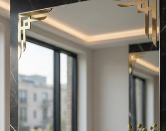 May include: A rectangular wall mirror featuring gold-toned metal corner accents with geometric, angular lines. The mirror is mounted on a dark, black marble-patterned surface, reflecting a bright interior room with windows and white flowers.