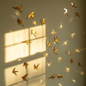 Puede incluir: Un conjunto de piezas de arte mural de pájaros de metal dorado en varias poses, montadas en una pared de color claro. Las aves están dispuestas para crear una sensación de vuelo, con sombras que añaden profundidad. La escena está iluminada por luz natural.