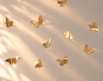 May include: A collection of ten gold-colored butterfly-shaped wall decals with a reflective, mirror-like finish. Each butterfly is depicted in a different flight position, creating a dynamic arrangement across the surface.