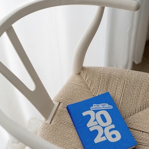 Peut inclure: Un livre de Sudoku bleu avec les chiffres "2026" en blanc, posé sur une chaise tressée. La chaise a un cadre en bois blanc. Le texte "Free 100+ Sudoku Puzzle" est également visible sur le livre.