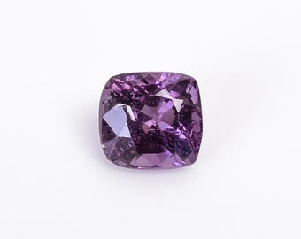 Può includere: Una gemma sfaccettata a taglio cuscino, di colore viola. La gemma ha una forma quadrata con angoli arrotondati e una superficie brillante e riflettente. Le sfaccettature creano un gioco di luci e ombre, esaltando la sua profondità e il colore. Su sfondo bianco.