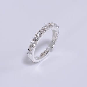 May include: A silver-colored ring with a row of clear, round gemstones. The ring is set on a white surface, and the gemstones sparkle. The ring is a jewelry item.