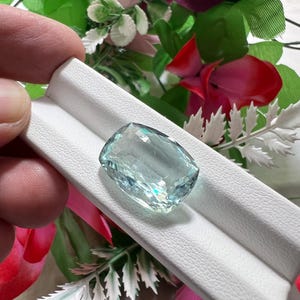Può includere: Una pietra preziosa acquamarina taglio a cuscino con una tonalità blu-verde pallido e una superficie sfaccettata, esposta su un espositore per gioielli bianco testurizzato. La gemma mostra un'elevata chiarezza e riflessione della luce.