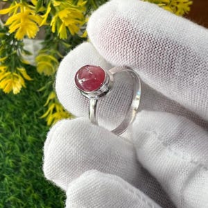 May include: A silver ring featuring a round, cabochon-cut, pink gemstone. The ring is held against a white background, with yellow flowers and green foliage visible in the background. The gemstone is approximately 0.5 inches in diameter.