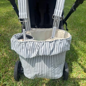 May include: A grey and white striped fabric stroller organizer bag featuring a ruffled top edge, an open main compartment, and two side bottle pockets. The bag attaches to the stroller handle with two adjustable fabric straps and silver metal clips.