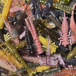 May include: A pile of colorful soft plastic fishing lures in various shapes, including worms, crawfish, and insect imitations. The lures are in shades of pink, yellow, green, and black, with some having glitter accents.