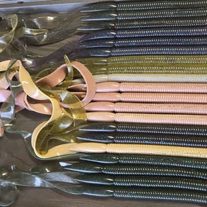 May include: An assortment of soft plastic fishing lures in various colors, including green, pink, and black, arranged on a metal tray. The lures have different shapes, including curly tails and ribbed bodies, designed for attracting fish.
