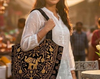 May include: A black velvet tote bag featuring intricate gold embroidery of an elephant surrounded by floral and vine patterns. The bag has a wide shoulder strap and a structured shape, showcasing detailed needlework in a metallic gold thread.