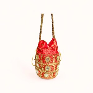 May include: A red, beaded drawstring bag with gold accents and a gold beaded handle. The bag features a pattern of gold embroidery and small, round mirrors. The top of the bag is gathered with a drawstring closure.