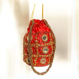 Puede incluir: Un bolso rojo de satén con adornos de cuentas doradas y detalles de espejo. El bolso presenta un asa trenzada de cuentas doradas y cordones dorados. El bolso está decorado con patrones florales dorados.