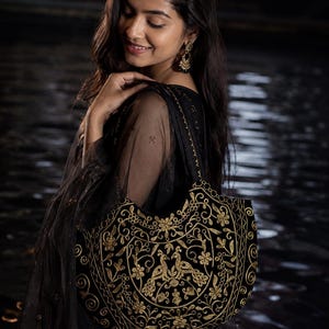 May include: A black shoulder bag featuring intricate gold thread embroidery in a floral and peacock motif. The bag has a curved top edge, two shoulder straps, and a structured shape, showcasing detailed needlework across the front panel.