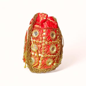 May include: A red drawstring bag with gold beaded embellishments and mirror accents. The bag features a gold beaded border and a gold beaded drawstring. The bag is made of a shiny red fabric.