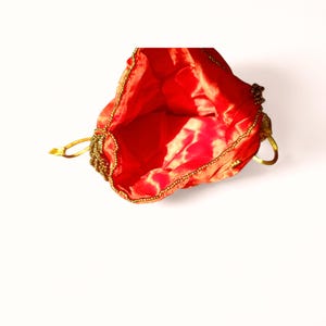 May include: A red satin drawstring bag with gold beaded trim and a gold drawstring. The interior of the bag is a lighter shade of red with a gold pattern. The bag is open, revealing the interior.