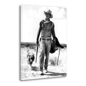May include: Monochrome canvas print depicting a cowboy in a desert setting, accompanied by a dog. The cowboy is dressed in a hat, shirt, and jeans, carrying a rifle and a saddle. The dog walks alongside him.