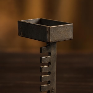 Könnte beinhalten: Ein handgefertigter Kerzenhalter aus dunklem Metall. Die rechteckige Oberseite wird von einem vertikalen Pfosten mit einer Reihe ausgeschnittener rechteckiger Formen getragen. Die Basis hat eine flache, quadratische Form. Der Kerzenhalter ist für eine einzelne Kerze konzipiert.
