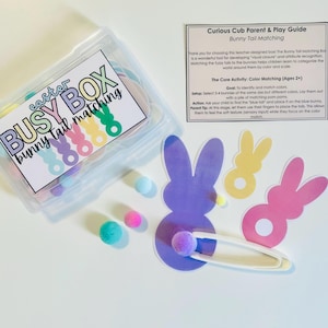 May include: An Easter-themed Busy Box with bunny tail matching game. The set includes a clear plastic container with colorful pom-poms, bunny cutouts, and a parent guide. The box is designed for color matching and visual closure activities.