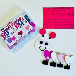 May include: A Valentine's Day Busy Box with a love bug name spelling theme. The box contains heart-shaped pieces in various colors. A paper love bug with googly eyes and heart-shaped segments spelling "PAPA" is also present.