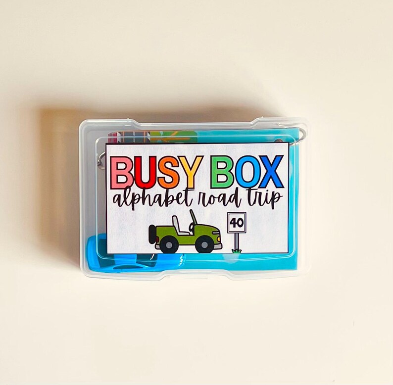 May include: A clear plastic box containing a colorful "Busy Box Alphabet Road Trip" set. The box includes a card with a green jeep illustration and a speed limit sign. The text on the card is in multiple colors.