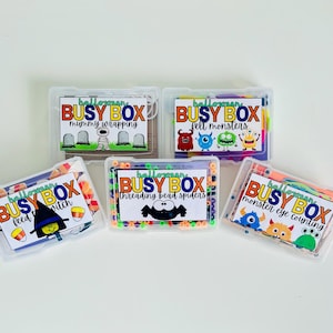 May include: Five clear plastic rectangular containers labeled Halloween Busy Box. The labels feature colorful text and graphics including a mummy, felt monsters, a witch, bead spiders, and monster eyes, with contents visible inside each box.