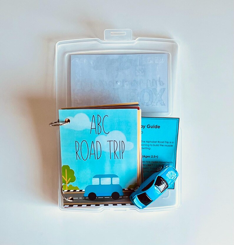 May include: A clear plastic case containing an educational toy set. The set includes a small booklet titled "ABC ROAD TRIP" with a blue car illustration, a blue toy car, and a guide. The booklet is bound with a metal ring.