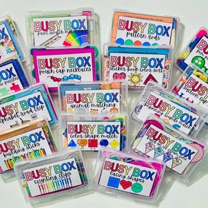 Op de afbeelding: Een verzameling doorzichtige Busy Boxes, elk gelabeld met educatieve activiteiten zoals namen spellen, patroonstaven en kleuren matchen. De dozen bevatten kleurrijk materiaal voor het leren en spelen van kinderen.