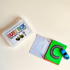 May include: A clear plastic box labeled "BUSY BOX alphabet road trip" with a green jeep illustration. An open book with a green page featuring a black road and the letter "C" and a blue toy car.