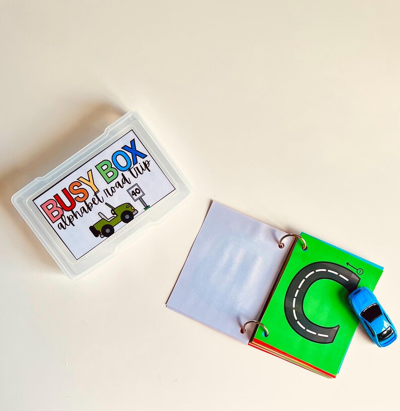 May include: A clear plastic box labeled "BUSY BOX alphabet road trip" with a green jeep illustration. An open book with a green page featuring a black road and the letter "C" and a blue toy car.