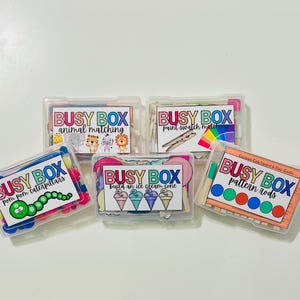 May include: Five clear plastic boxes labeled "BUSY BOX" with different activities: animal matching, pom pom caterpillars, paint swatch matching, build an ice cream cone, and pattern rods. Each box contains colorful craft supplies and printed cards.