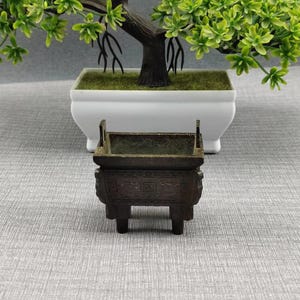 May include: A small, rectangular, dark bronze-colored metal incense burner featuring four short legs and intricate relief patterns on its exterior sides. The vessel has two upright handles on the top rim and an open, hollow interior space.