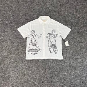 Puede incluir: Camisa blanca con botones y mangas cortas. La camisa presenta un dibujo lineal negro de dos figuras con vestimenta tradicional. Tiene cuello y una etiqueta adjunta en la parte inferior derecha.