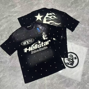 May include: Black t-shirts with white star and flame graphics. One shirt features the text "Hellstar" with a star and flame design. The shirts have a speckled pattern and are displayed with a clear plastic bag with a matching logo.