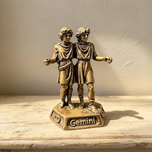 Op de afbeelding: Een bronskleurig metalen beeldje met twee figuren die naast elkaar op een zeshoekige voet staan. De voet is voorzien van het in reliëf aangebrachte opschrift Gemini. De figuren dragen gedrapeerde tunieken en sandalen.