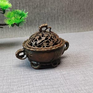 Puede incluir: Un quemador de incienso de bronce oscuro con un cuenco redondo, dos asas laterales y una tapa extraíble. La tapa presenta un intrincado patrón calado de hojas de arce, mientras que el cuenco muestra tallos y hojas de bambú en relieve.