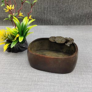 May include: An oval-shaped dark bronze bowl featuring a decorative relief of three lily pads and a small fish on the rim. The bowl has a smooth, matte finish and a deep basin, providing a functional container for small items or decorative display.