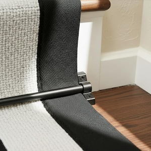 May include: A close-up of a stair runner with a black and white striped pattern. The runner is held in place by a black metal rod and brackets, attached to a wooden handrail. The background includes a white wall and dark wood flooring.
