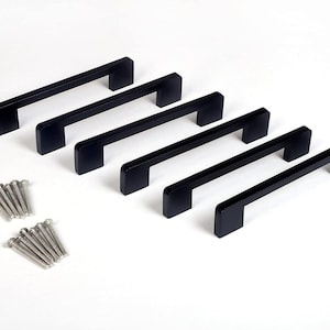 Könnte beinhalten: Sechs schwarze Schrankgriffe mit passenden Schrauben sind auf weißem Hintergrund angeordnet. Die Griffe haben ein schlichtes, modernes Design. Die Hardware ist für Schubladen oder Schränke geeignet.