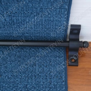 May include: A black curtain rod with a decorative bracket, mounted on a wooden surface. The rod is positioned in front of a blue textured fabric, creating a contrast of colors and textures. The bracket is attached to the wooden surface.
