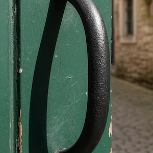 以下が含まれることがあります： 風化した緑色のドアにある、黒い装飾的なドアノッカーのクローズアップ。ノッカーはテクスチャード加工された仕上げと装飾的なデザインです。背景にはレンガ造りの建物と石畳の通りが写っています。