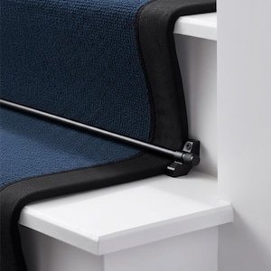 May include: A navy blue carpet runner with a black border, installed on a white staircase. A black metal rod secures the carpet. The carpet has a textured appearance. The image shows a close-up of the stairs.