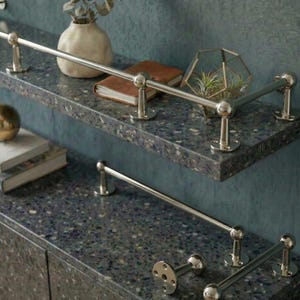 May include: A close-up of a modern bathroom shelf with a dark speckled countertop and brushed nickel towel bars. The shelf is decorated with a vase, books, and a geometric terrarium with plants. The wall is a muted teal color.