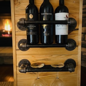 Può includere: Portabottiglie in legno con tre bottiglie di vino e due bicchieri. Il portabottiglie è realizzato in legno chiaro e tubi di metallo nero. Le bottiglie di vino sono scure con varie etichette.