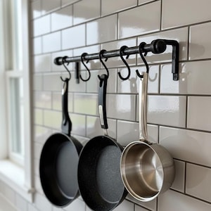 Op de afbeelding: Een zwarte metalen pannenrek met drie hangende potten en pannen. Het rek is bevestigd aan een witte betegelde muur. De potten omvatten twee zwarte koekenpannen en een roestvrijstalen steelpan.