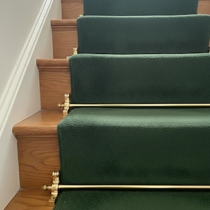 May include: A staircase with wooden steps and dark green carpet runners. The carpet is secured with gold-colored rods and decorative brackets. The steps are made of light brown wood, creating a contrast with the carpet.