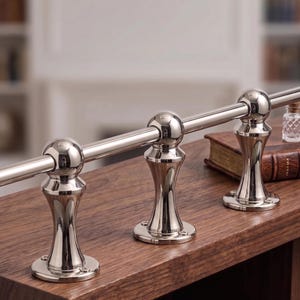 May include: A polished silver-tone metal railing system featuring three vertical supports with circular bases and spherical connectors. The horizontal rod passes through the center of each sphere, creating a continuous, reflective barrier structure.
