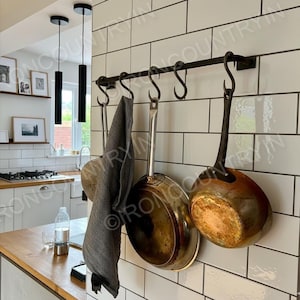 May include: A black wrought iron pot rack with six S-hooks, mounted on a white tiled wall. Two copper-bottomed pans, a stainless steel pan, and a gray towel hang from the hooks. The kitchen setting includes a wooden countertop and black pendant lights.