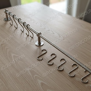 May include: A stainless steel kitchen rail with several S-hooks, designed for hanging utensils. The rail is mounted on the wall with circular brackets. Additional S-hooks are placed below the rail. The overall design is modern and functional.