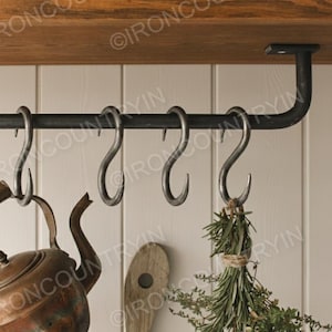Puede incluir: Un estante de metal negro para ollas con seis ganchos en forma de S, montado debajo de un estante de madera. Una tetera de cobre, una tabla de cortar de madera y un manojo de hierbas cuelgan de los ganchos. Decoración de cocina rústica.
